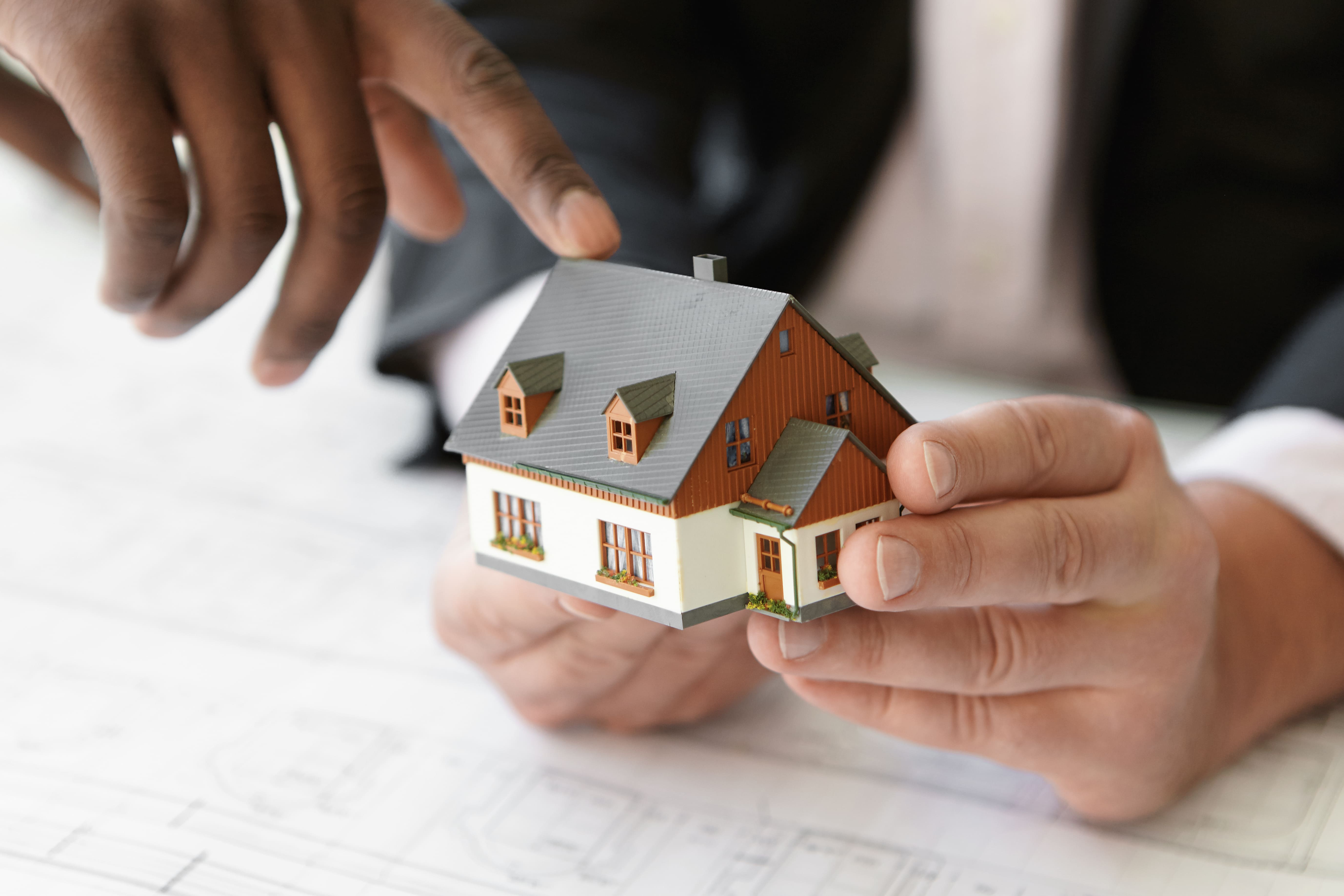 Two people holding and discussing a miniature house model over property plans, representing real estate consultation or home buying decision
